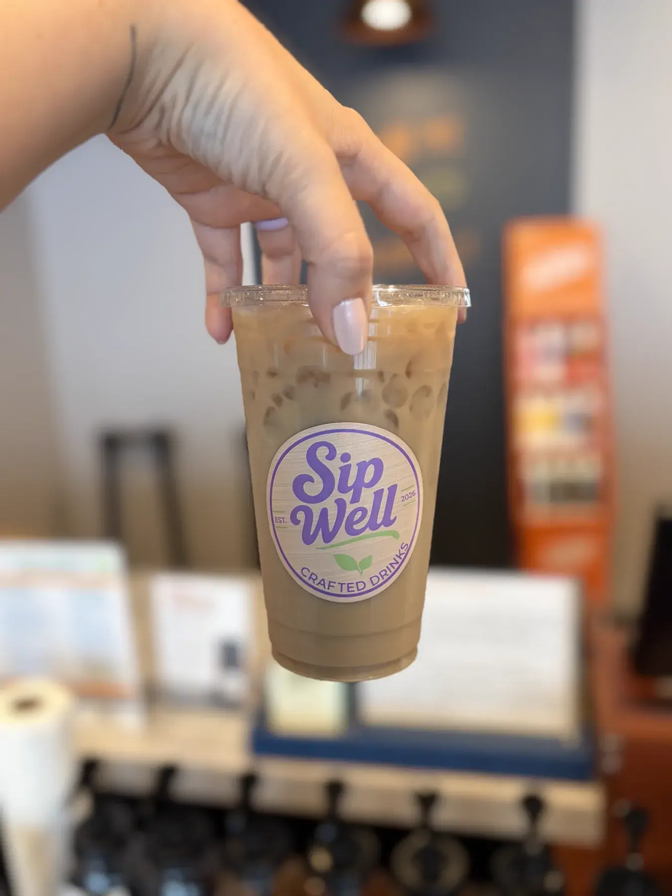 A hand holding a Sip Well cup filled with a cream-coloured iced drink