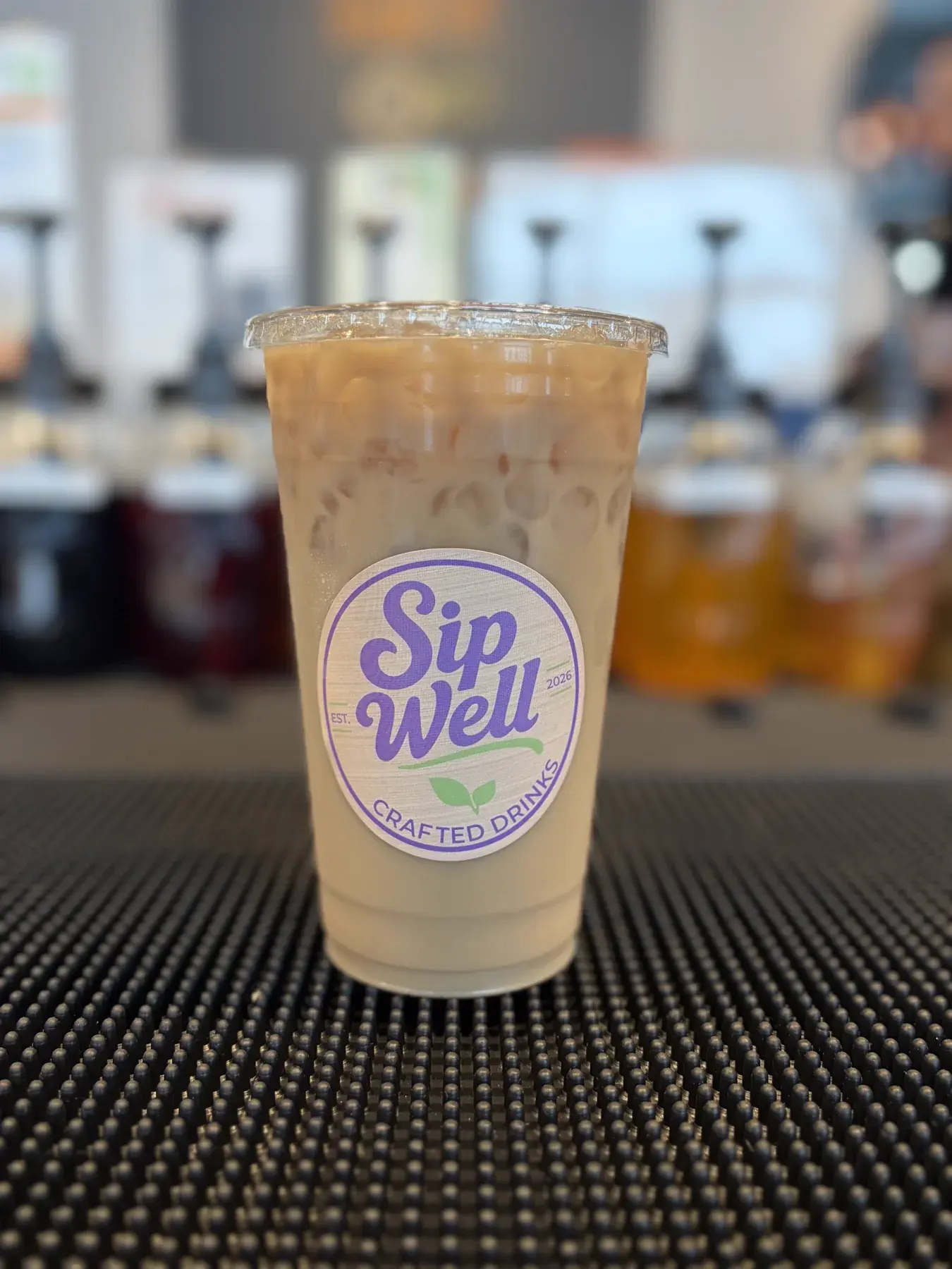 A Sip Well cup of creamy iced soda on the counter mat with the syrup pump jars behind it
